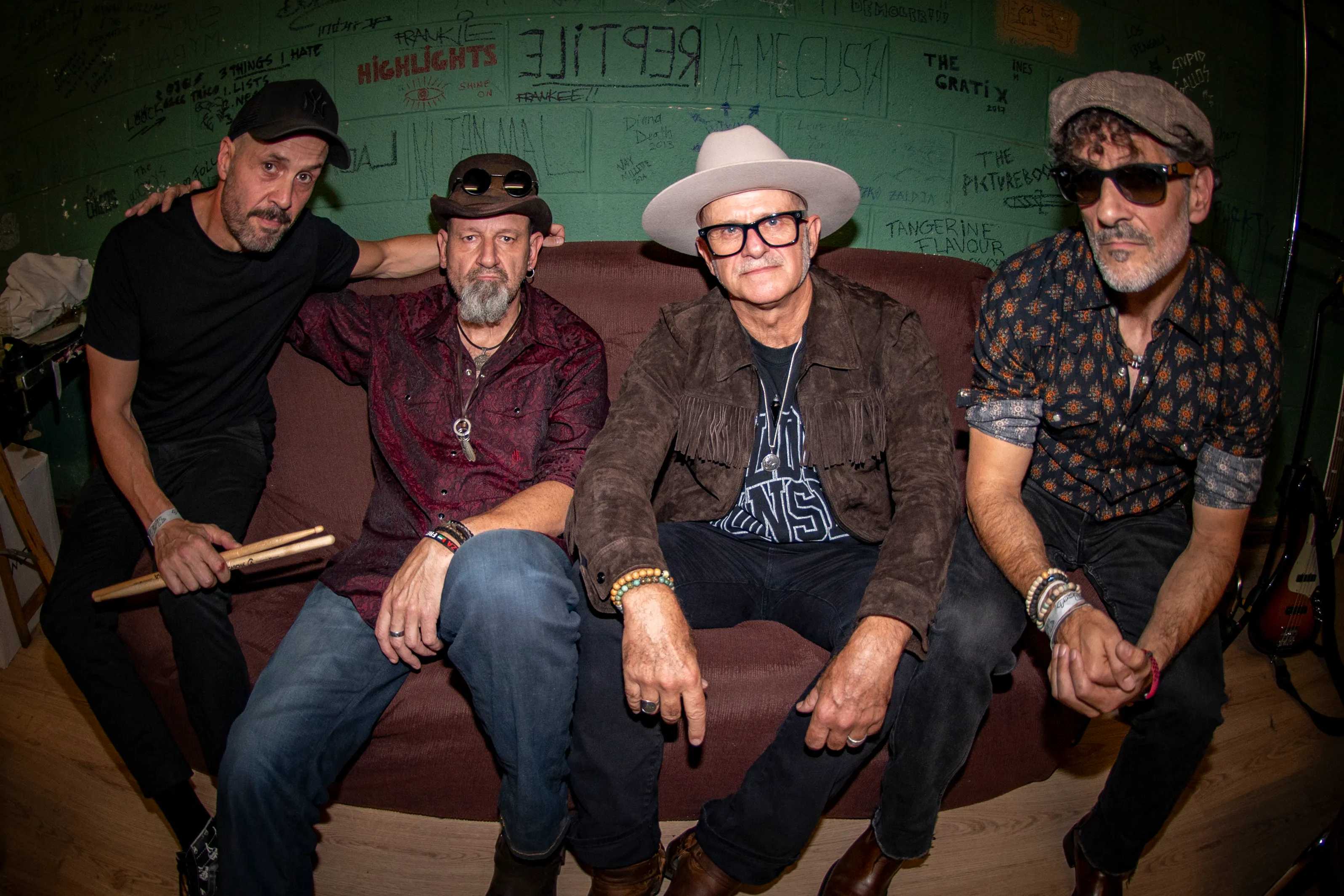 The four band members on a sofa backstage, in front of a green wall covered in graffiti