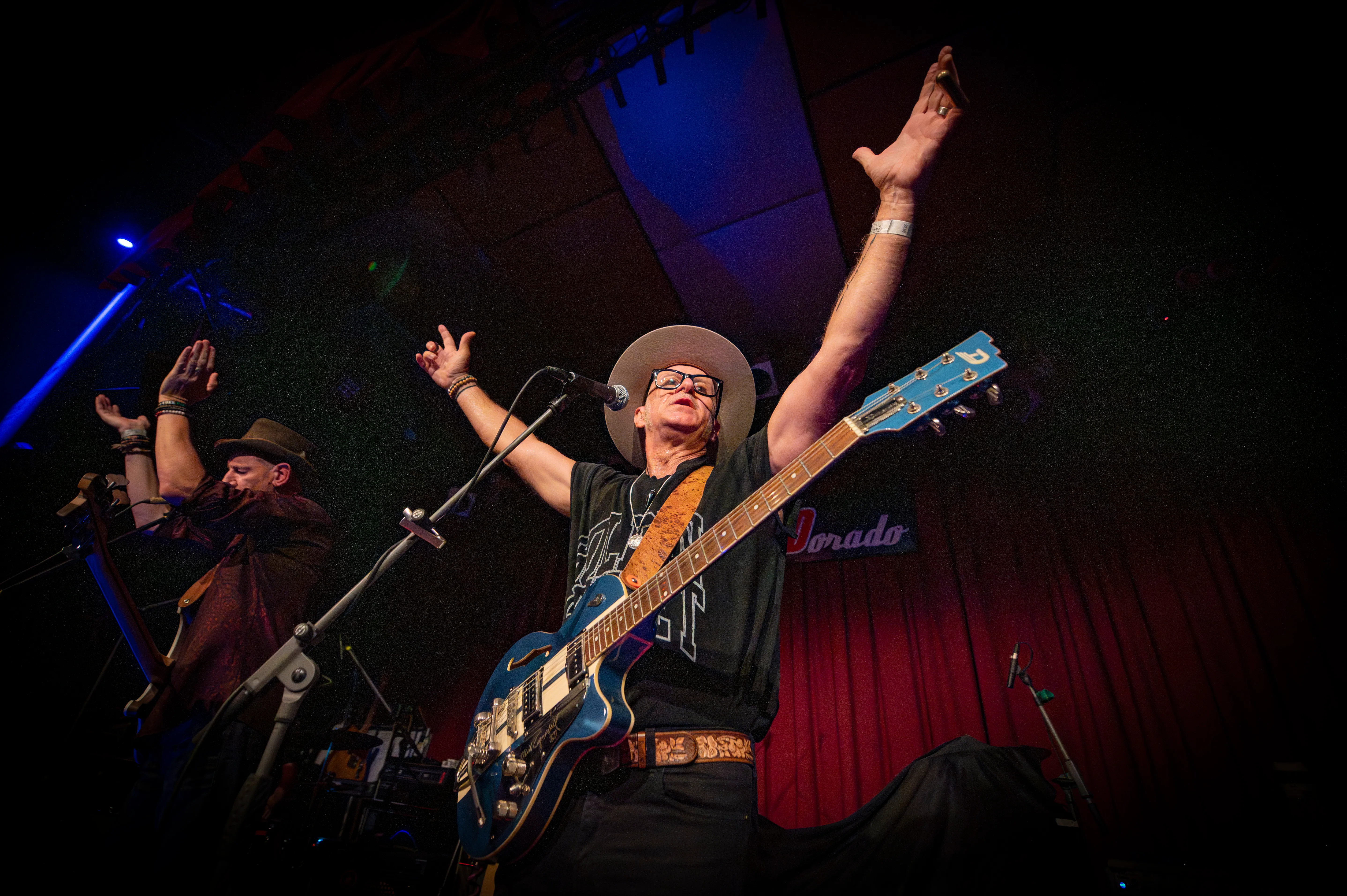The frontman on stage with a blue guitar and one arm raised, his bandmates applauding
