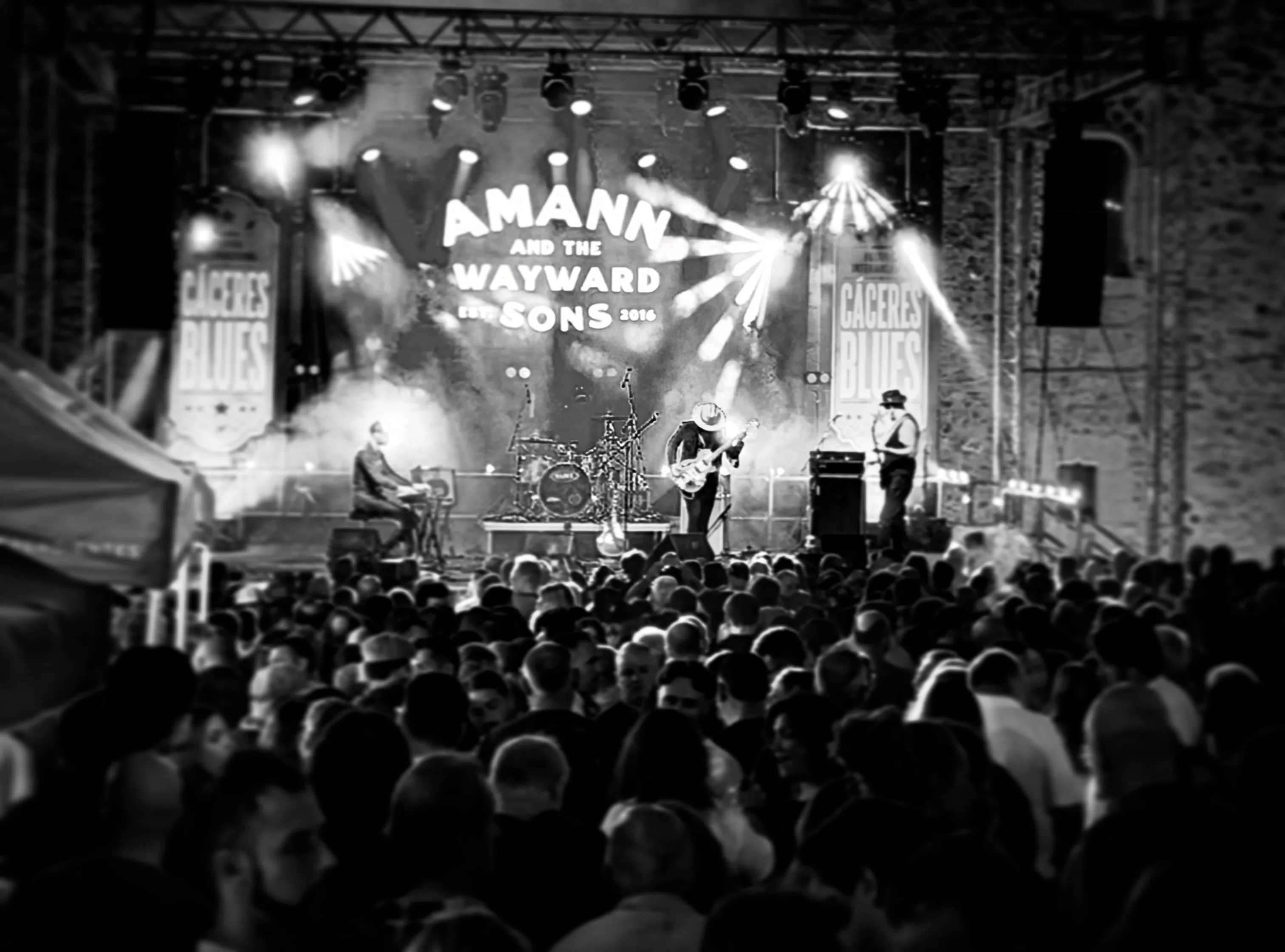 The band on an open-air festival stage under its own logo, seen over a packed crowd, in black and white