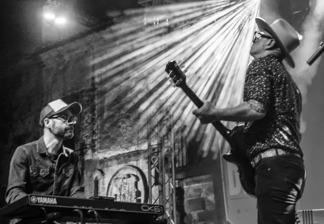The singer playing guitar under a burst of stage lights, the keyboard player beside him, in black and white