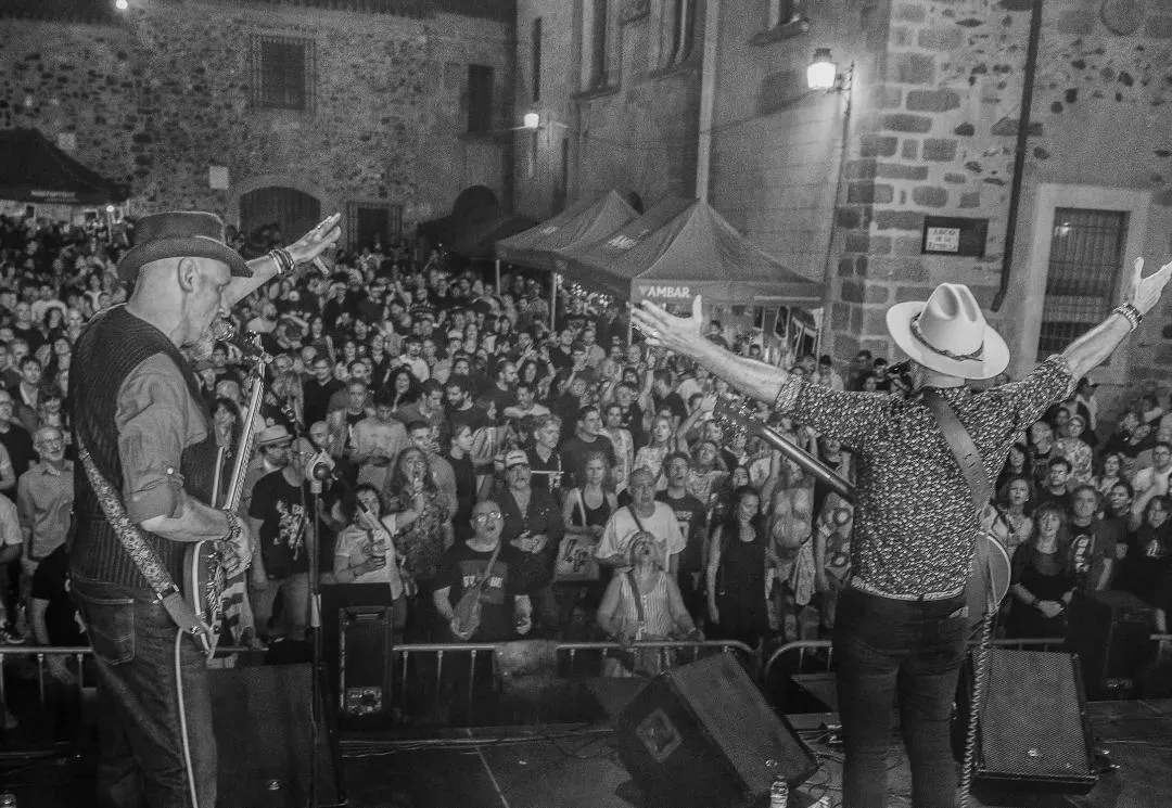 The band seen from behind, arms raised to a packed square, in black and white